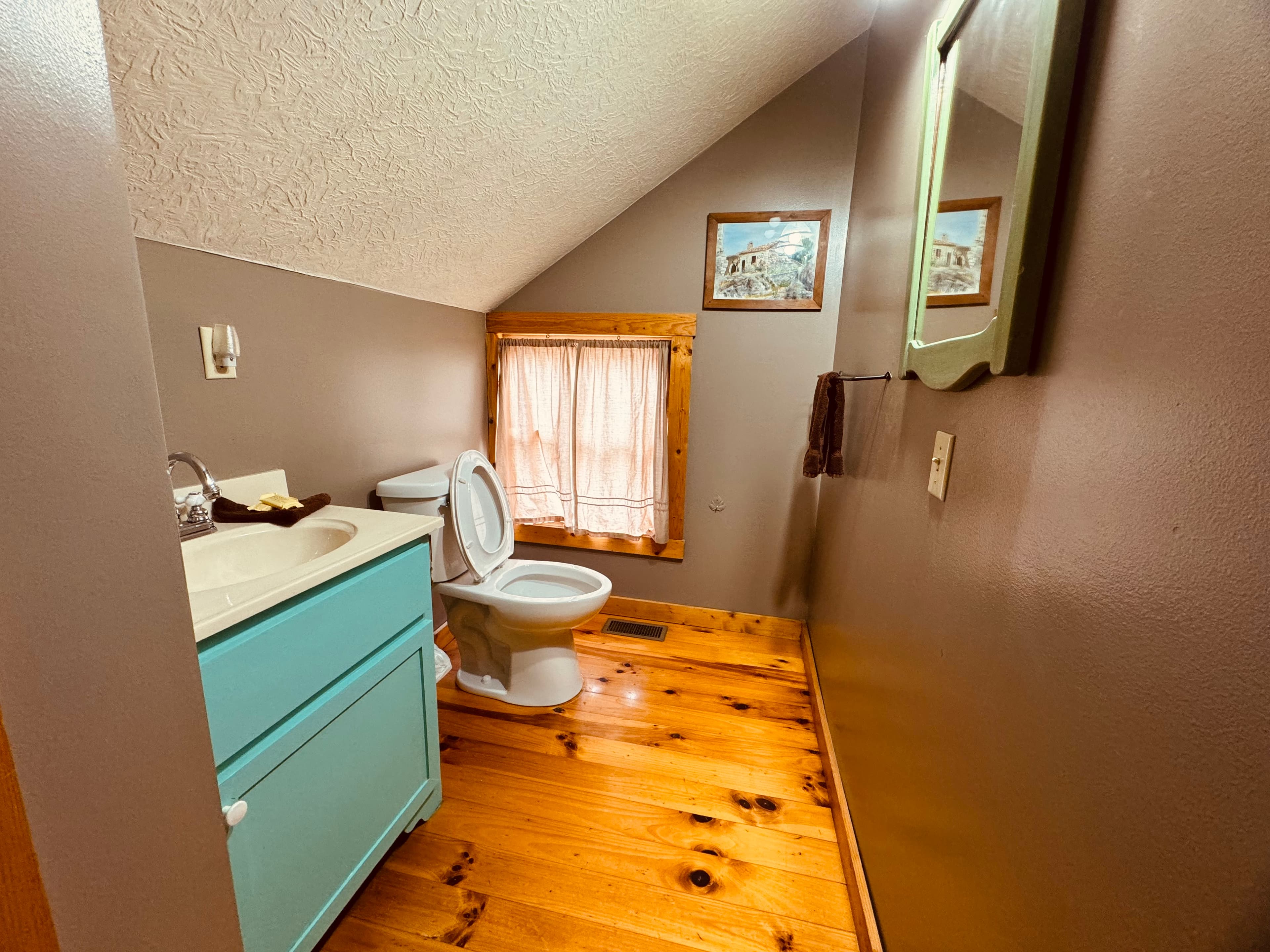 A small bathroom featuring a blue vanity, white toilet, and wooden flooring under angled ceilings.