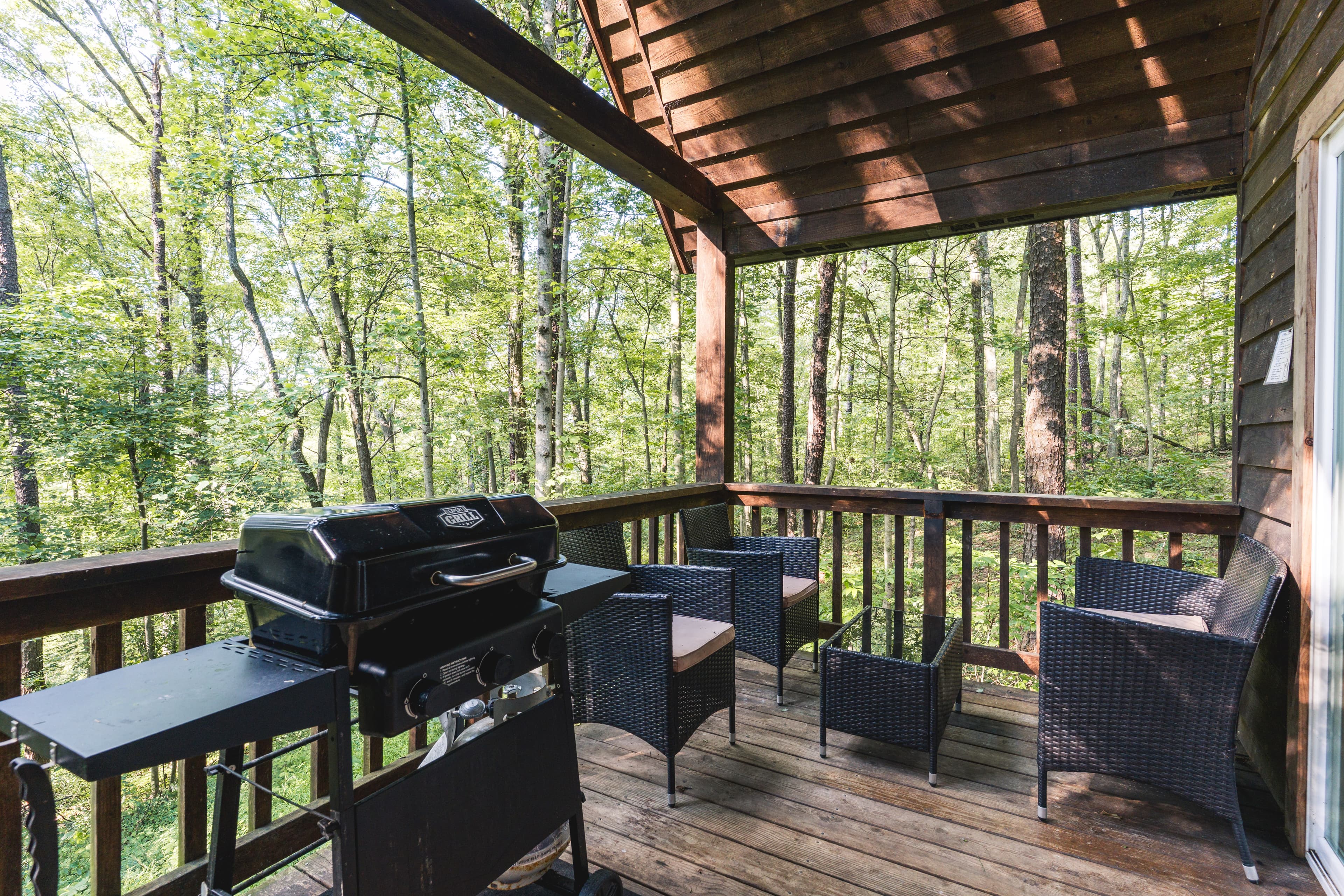 A wooden deck surrounded by trees features a barbecue grill and wicker chairs.