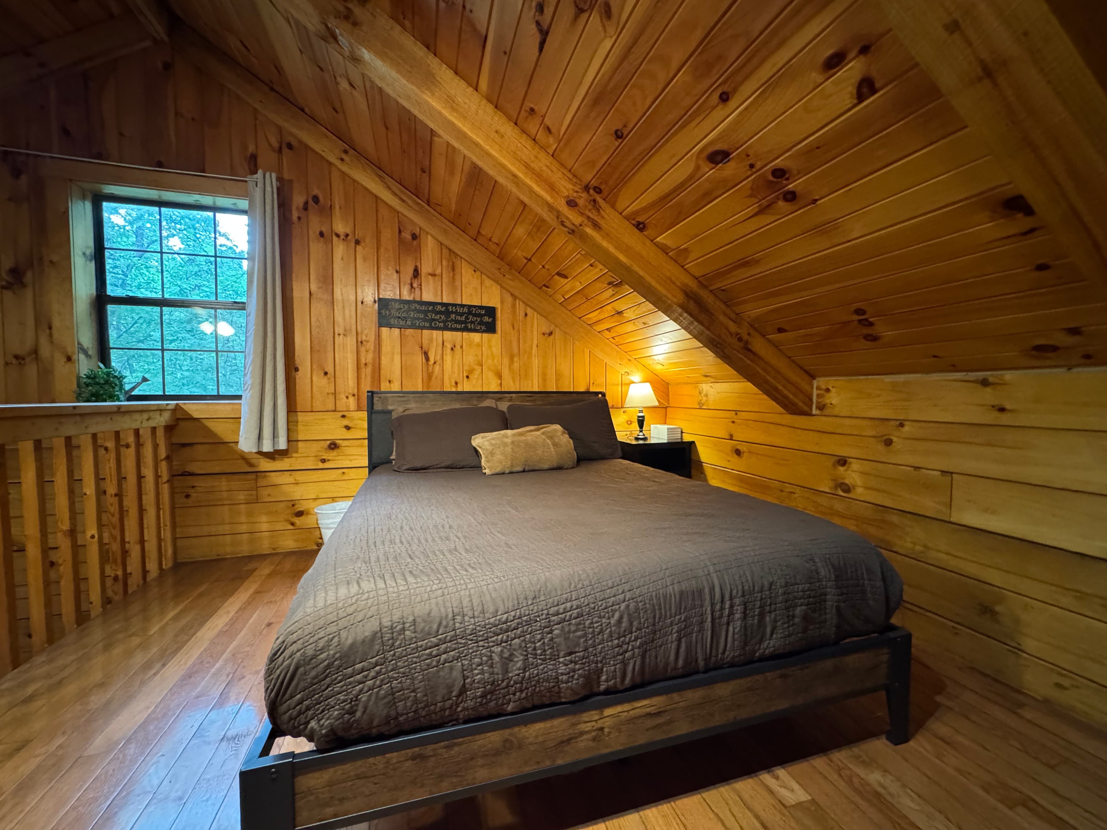 A cozy wooden bedroom with a bed, nightstand, and a window letting in natural light.