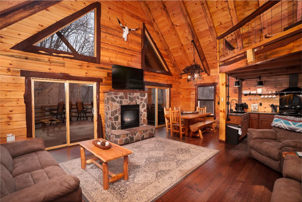 Cozy wooden cabin interior featuring a stone fireplace, large windows, and rustic furnishings.