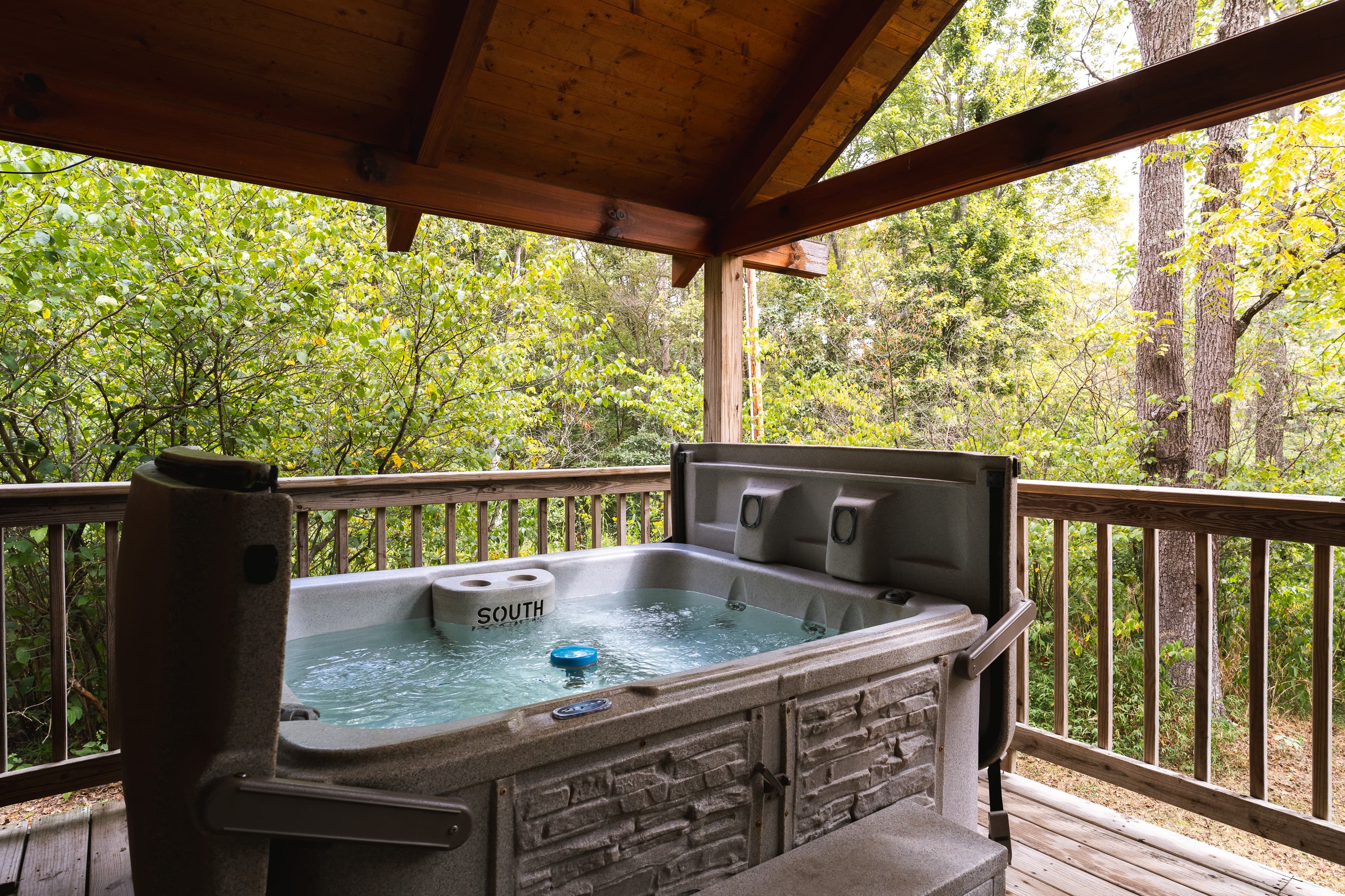 A hot tub sits on a wooden deck surrounded by lush greenery and a wooden roof overhead.