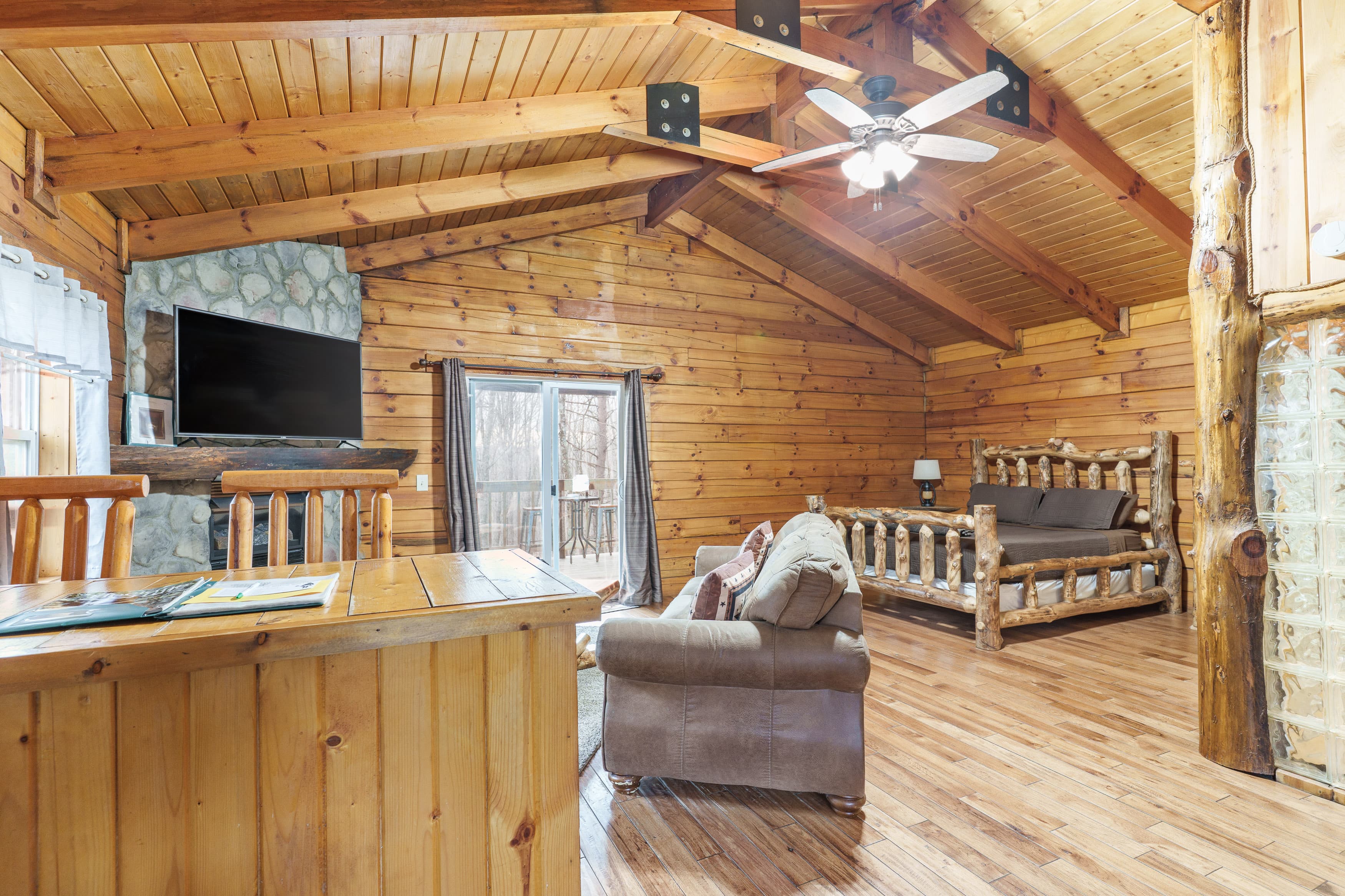Cozy wooden living area featuring a large window, rustic furniture, and a television mounted on a stone wall.
