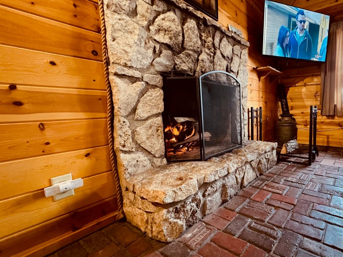 A cozy stone fireplace with a fire burning, set in a wooden cabin, alongside a television displaying a scene.