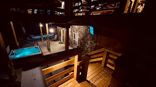 A wooden deck overlooks a stone house with a hot tub and outdoor lighting at night.