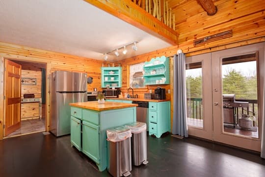 A cozy kitchen with turquoise cabinets, wooden walls, and large windows leading to a deck.