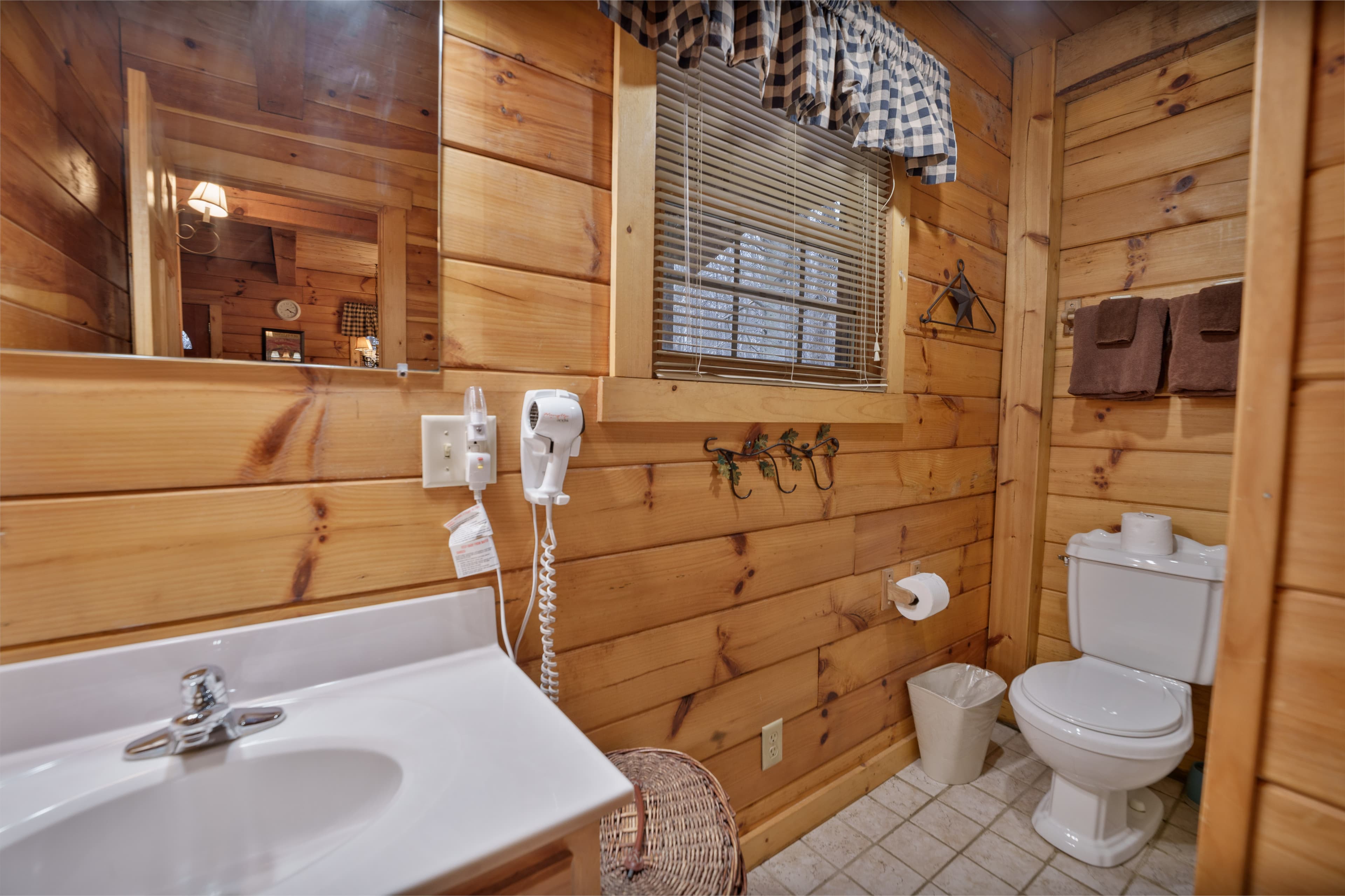 A rustic wooden bathroom featuring a sink, toilet, and window with checkered curtains.