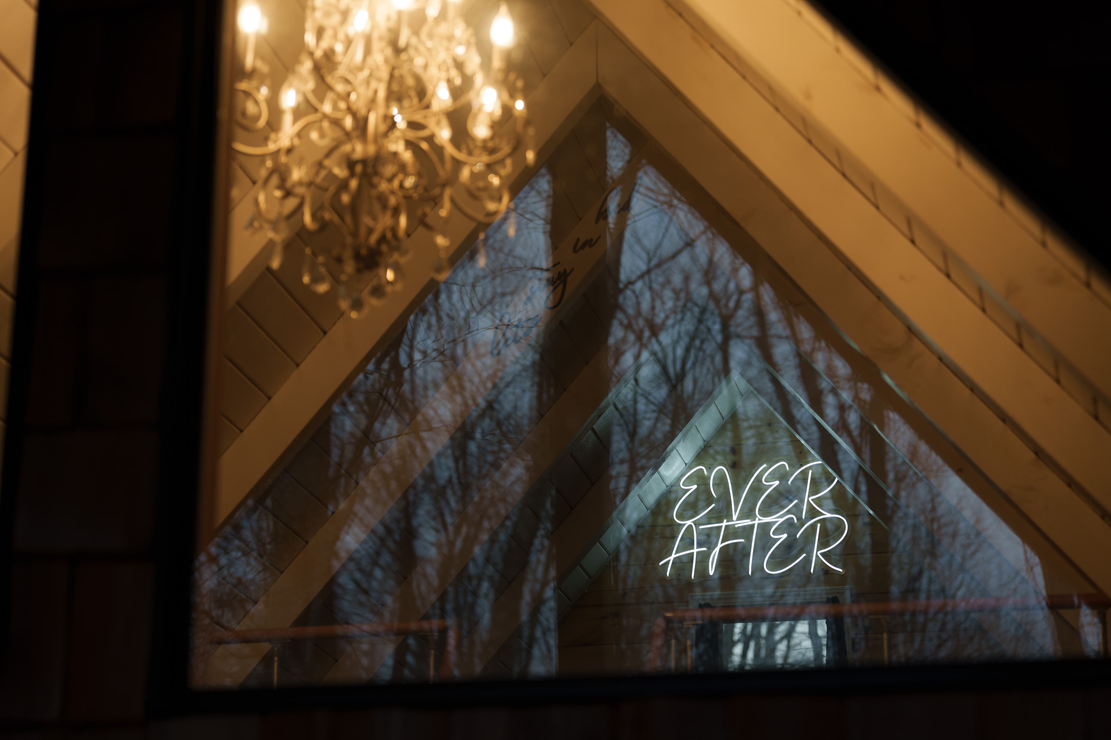 A glowing "EVER AFTER" neon sign is reflected in a window beneath a chandelier inside a wooden structure, with trees visible outside.