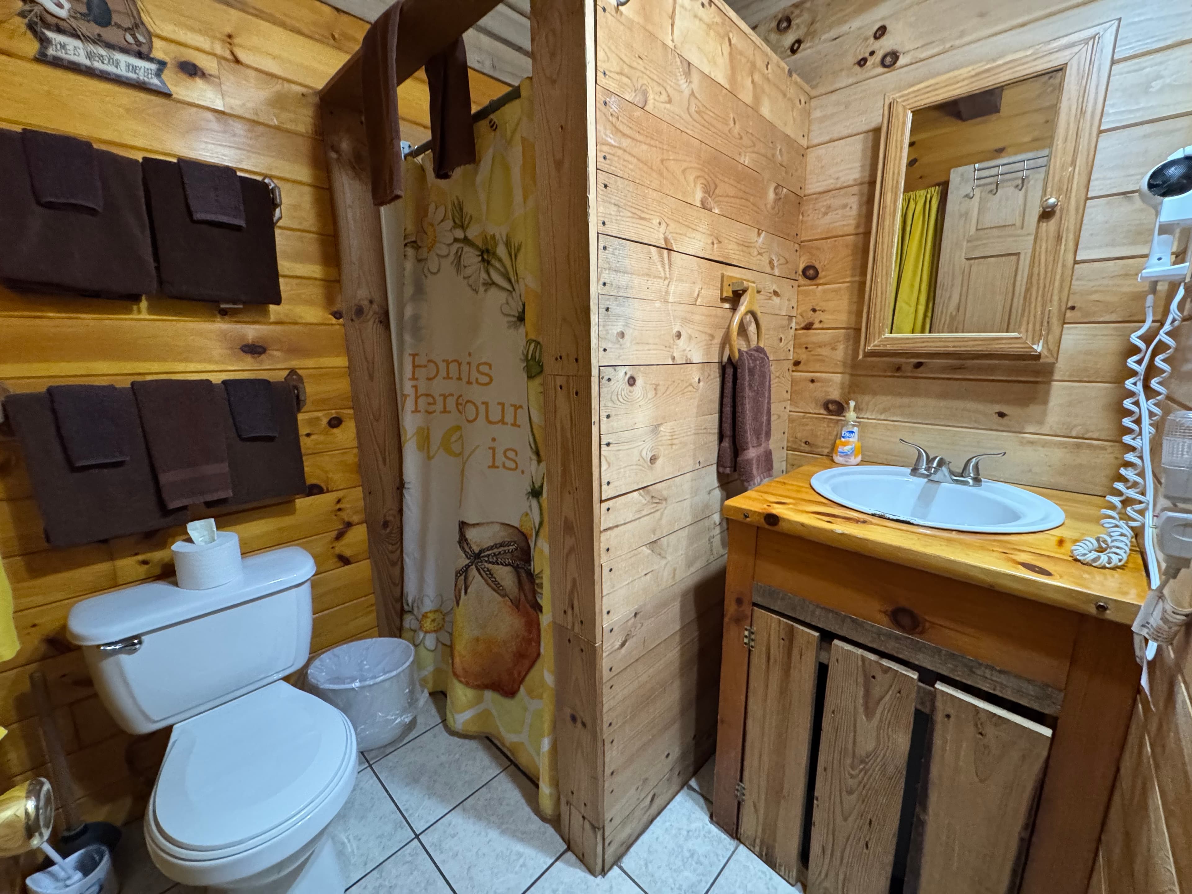 A cozy wooden bathroom featuring a shower, sink, and toilet, with brown towels and a yellow shower curtain.