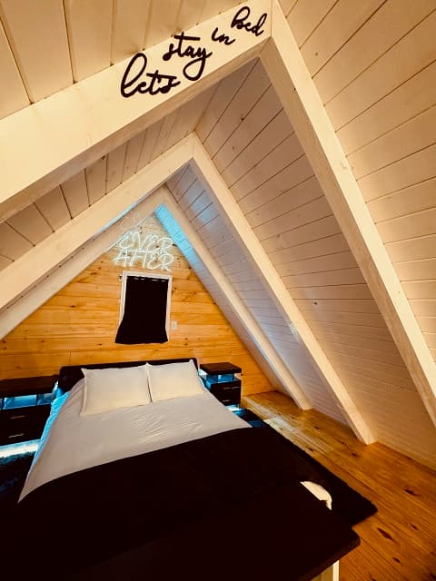 A cozy attic bedroom featuring a bed with white linens, wooden walls, and a neon sign reading "Happily Ever After."