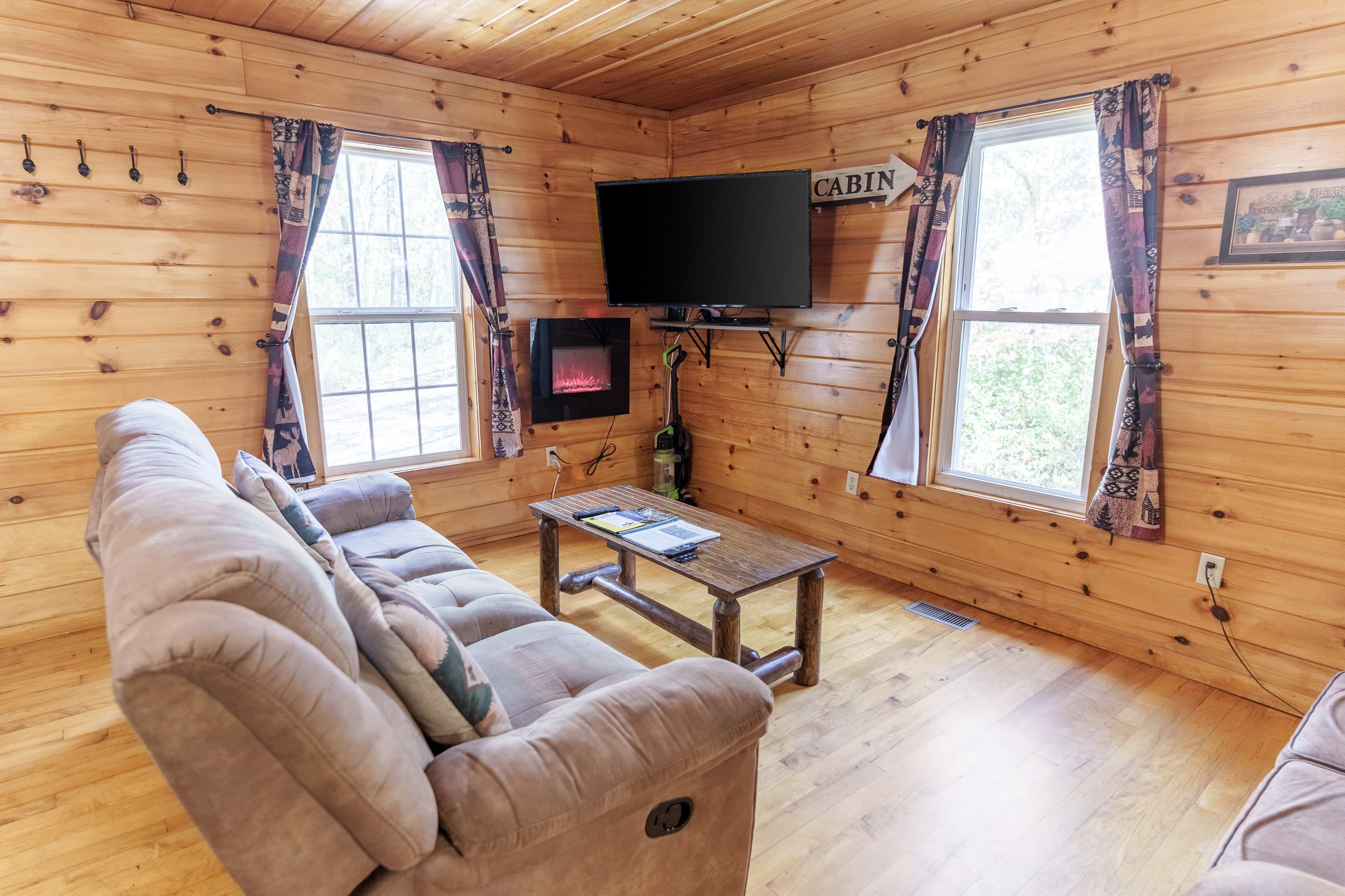 A cozy cabin living room featuring a sofa, wooden coffee table, and a mounted TV beside large windows.