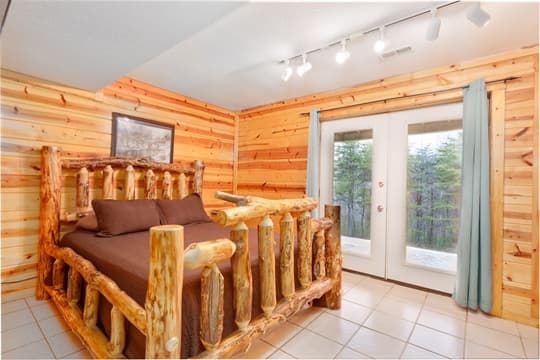 A rustic bedroom featuring a wooden log bed, tile floor, and large glass doors leading to a forested view.