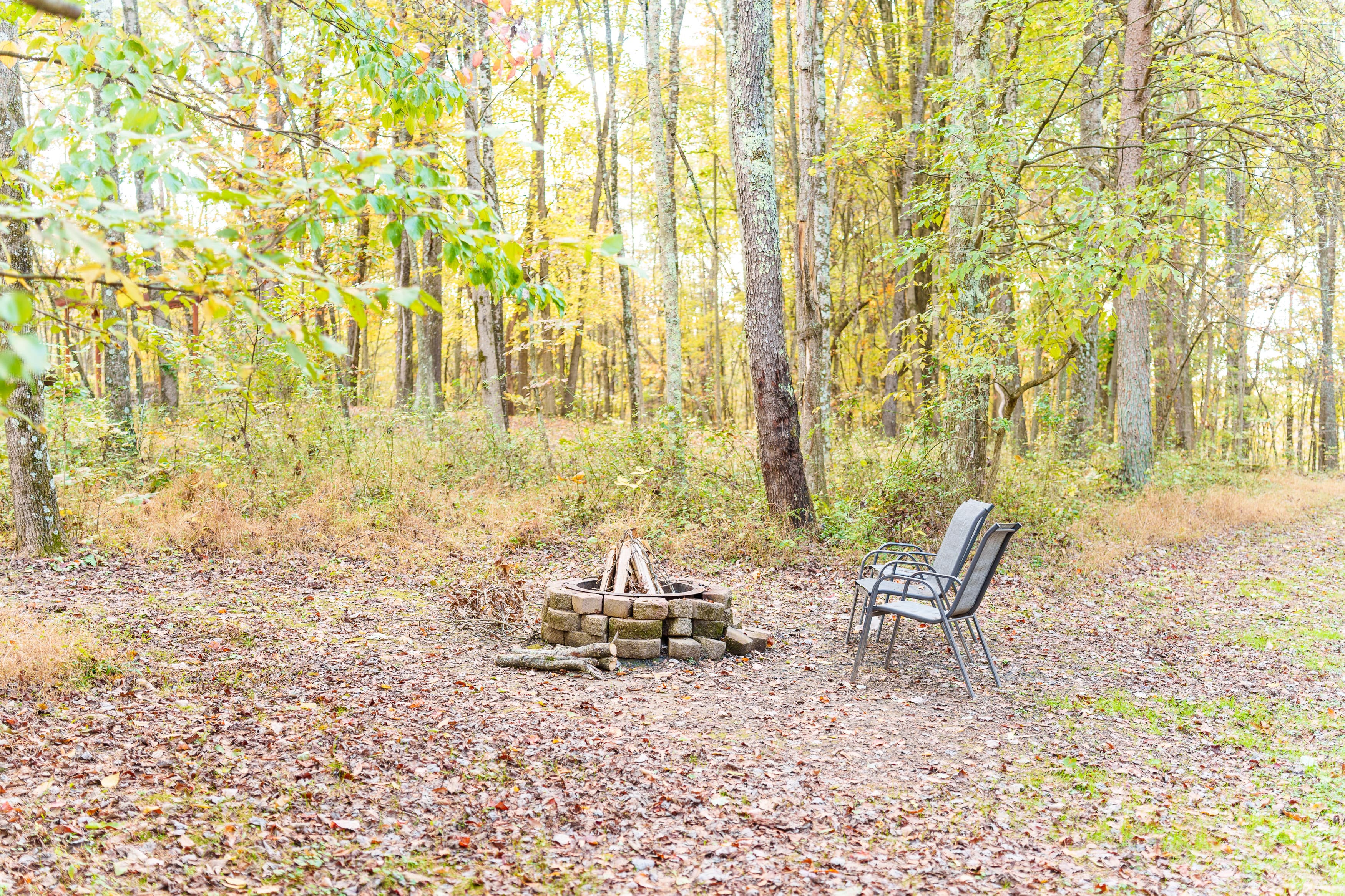 A rustic campfire pit surrounded by trees, with a pair of chairs nearby.