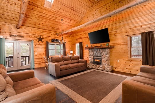 A cozy log cabin living room featuring a stone fireplace, large windows, and a mounted TV.