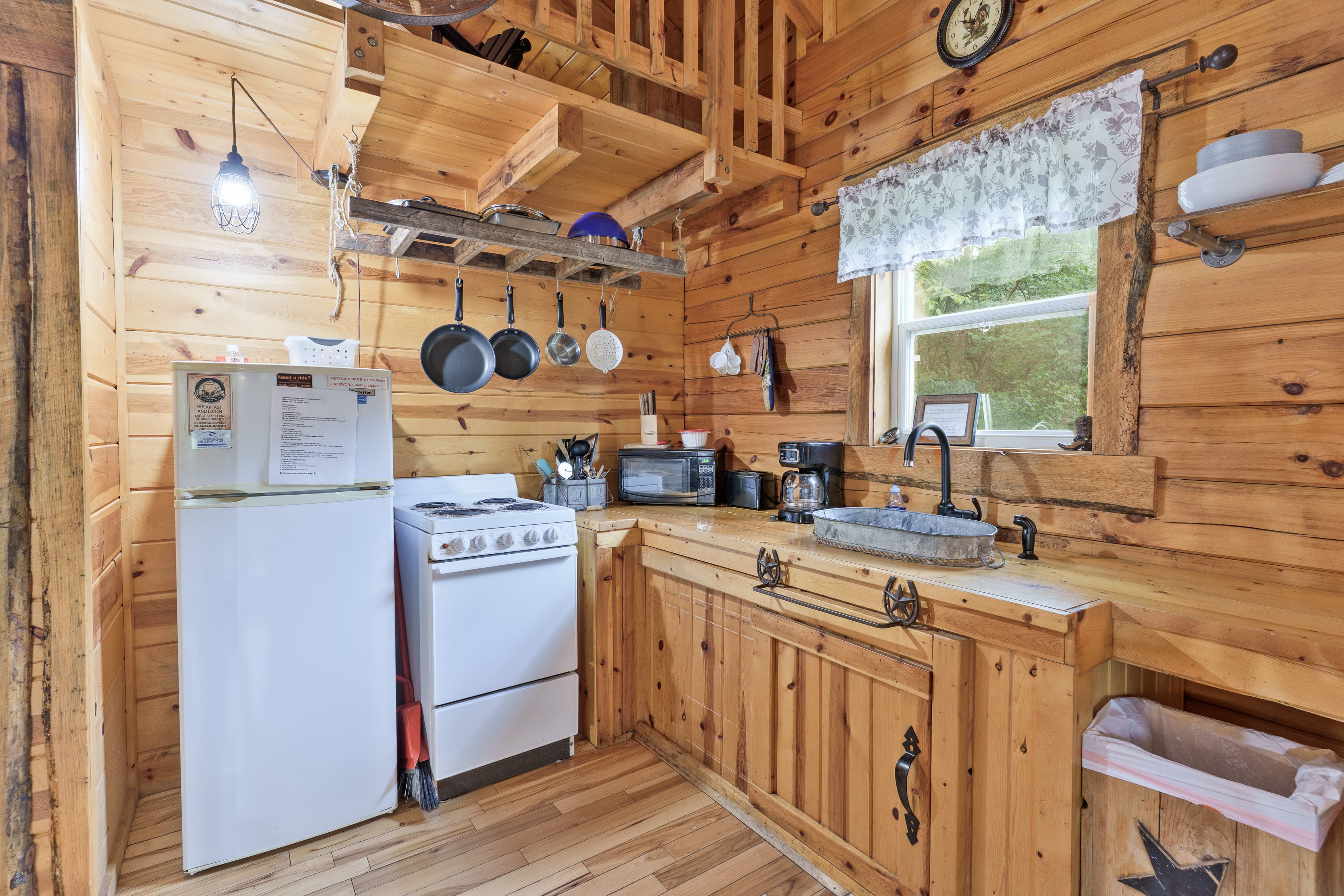 A cozy wooden kitchen with a stove, fridge, and rustic cabinetry, featuring hanging pots and a window with floral curtains.