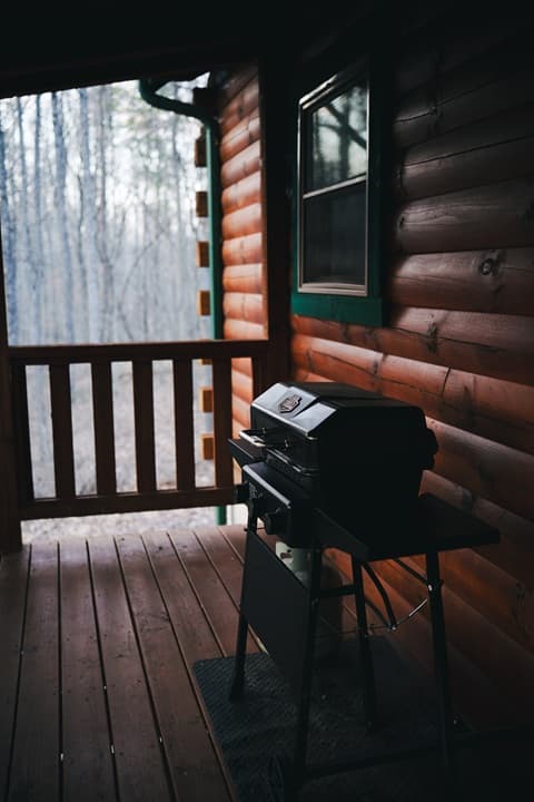 A grill sits on a wooden porch with views of a forest in the background.