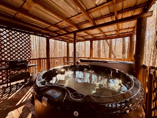 A hot tub sits on a wooden deck surrounded by trees under a covered structure.