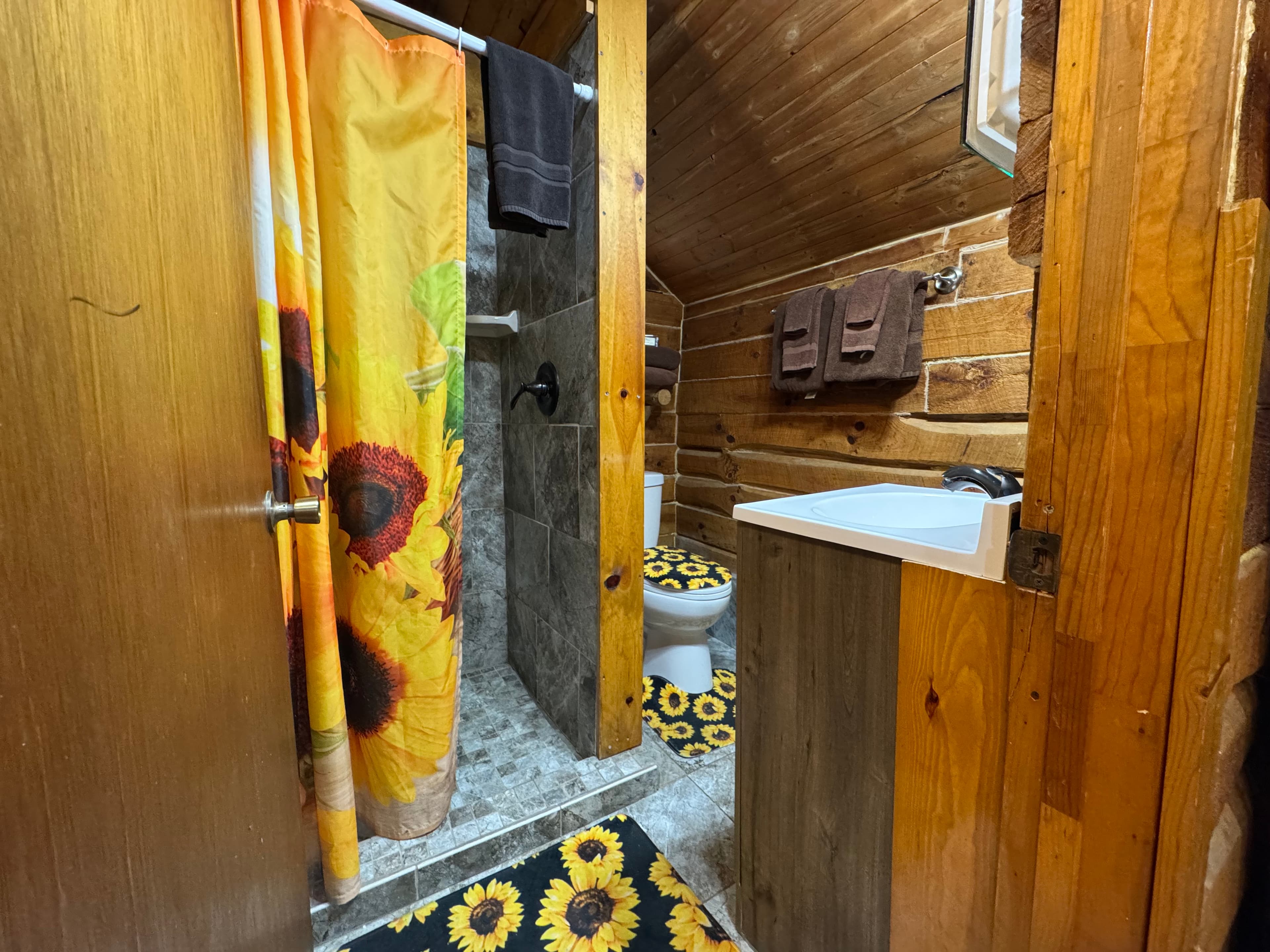 A rustic bathroom featuring a sunflower shower curtain, patterned rug, and wooden walls.