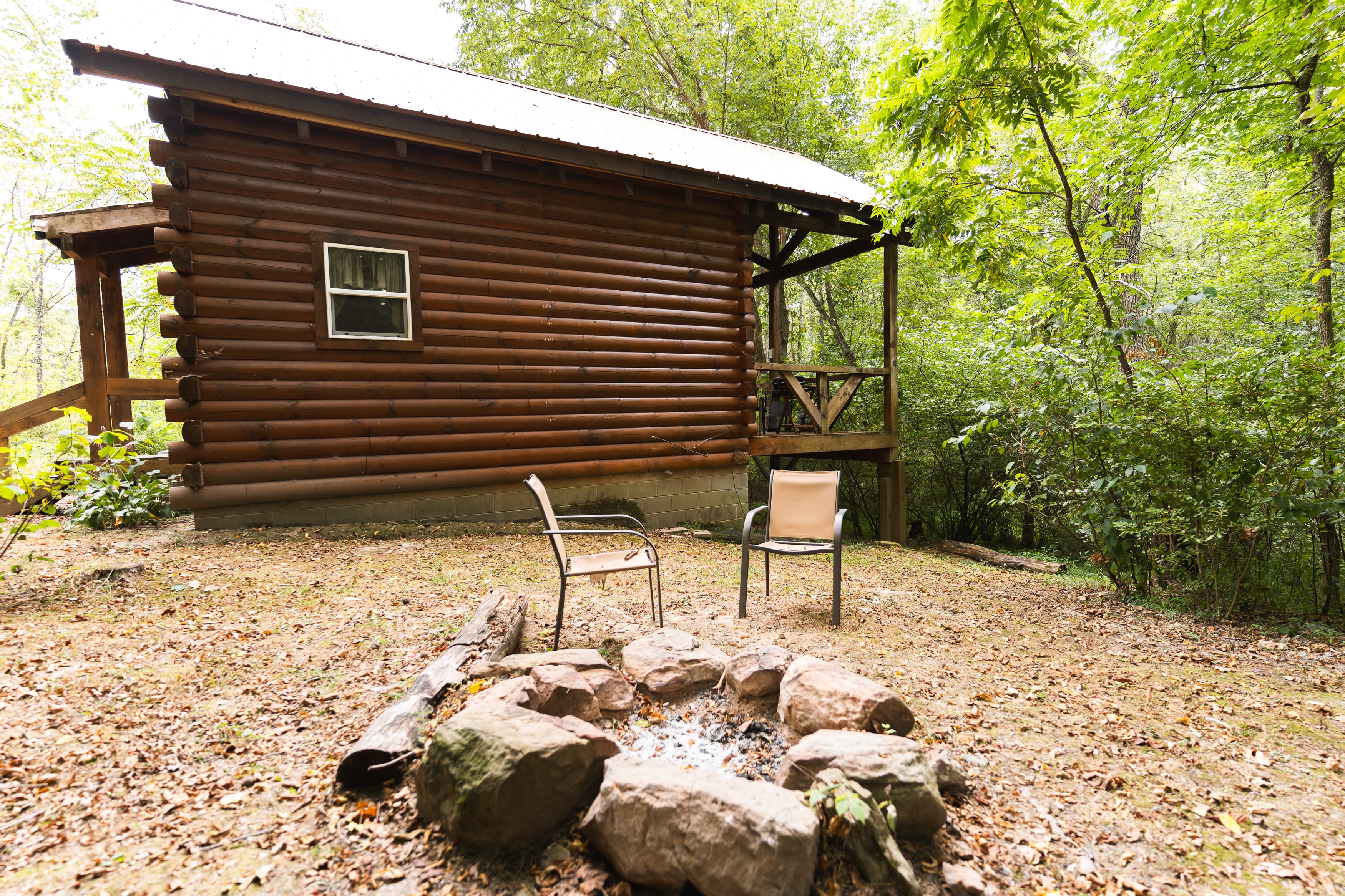 A rustic log cabin with a fire pit and two chairs set in a wooded area.