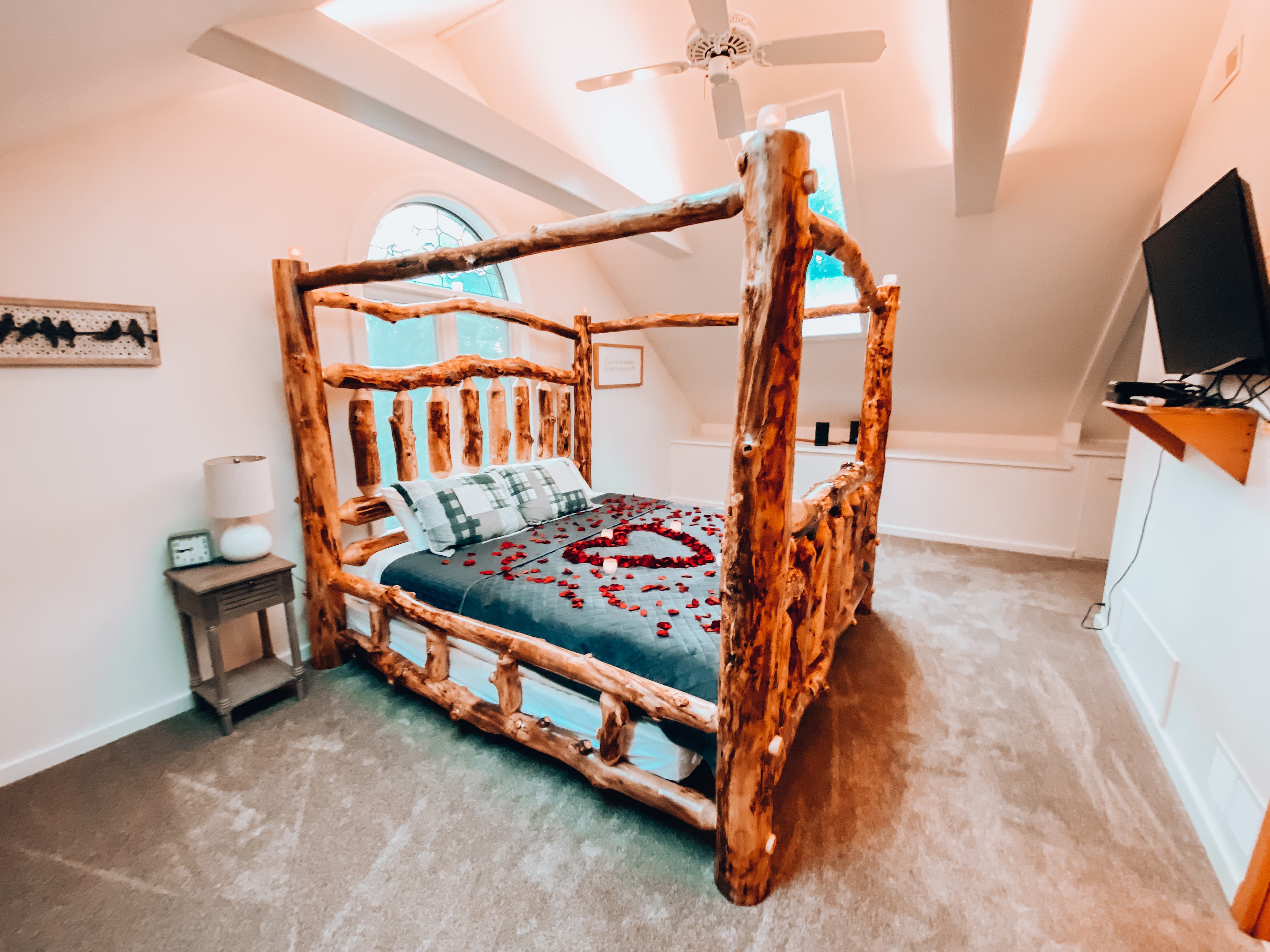 Rustic wooden bed adorned with rose petals in a cozy, well-lit attic bedroom.
