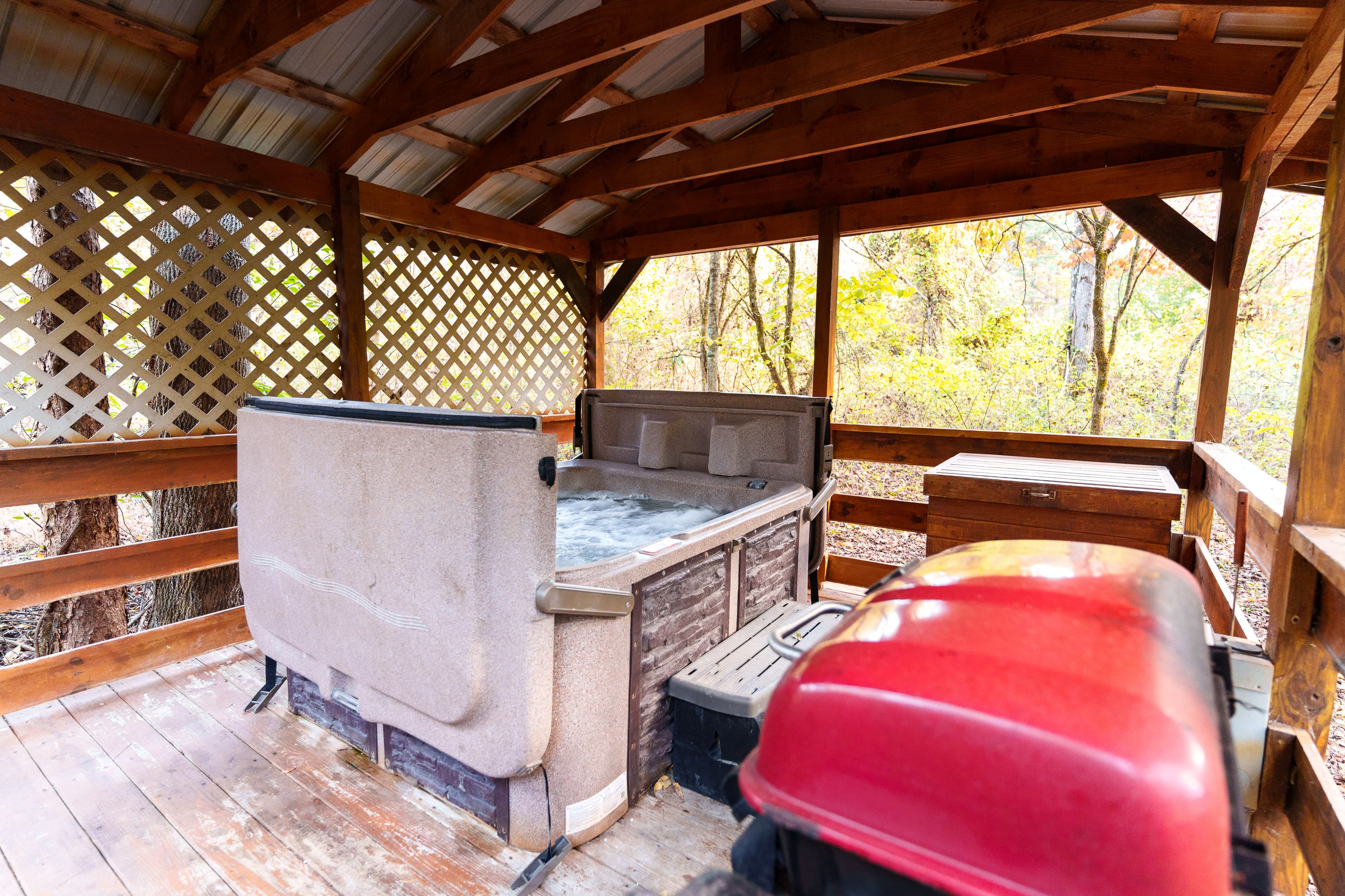 A wooden gazebo with a hot tub and a red tool chest inside, surrounded by trees.