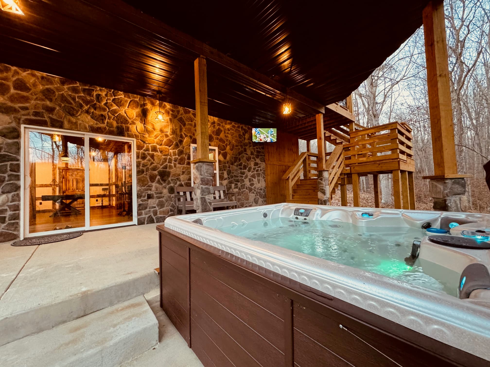 A hot tub is placed on a covered patio next to a stone wall, with wooden stairs leading to an upper level and a TV mounted nearby.
