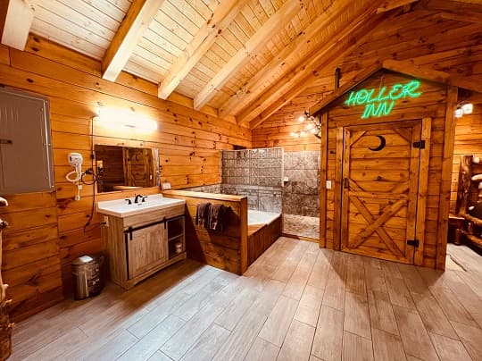 A rustic bathroom featuring wooden walls, a sink area, a large bathtub, and a neon "Holler Inn" sign.