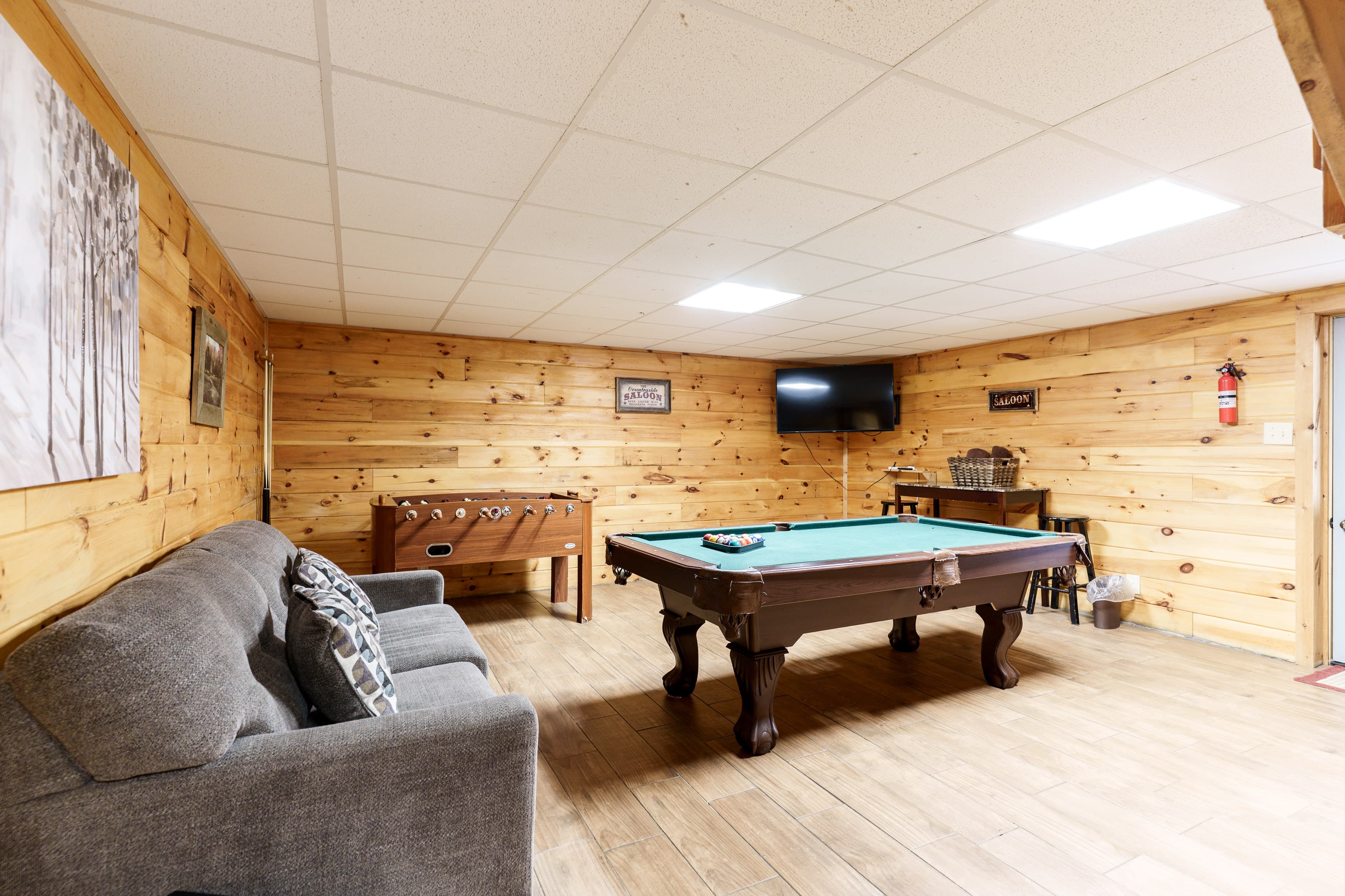 A cozy basement game room featuring a pool table, foosball table, and a gray couch against wooden walls.