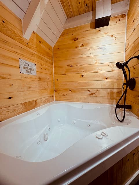A cozy, wooden-paneled bathroom featuring a white jacuzzi tub and a rustic charm sign.