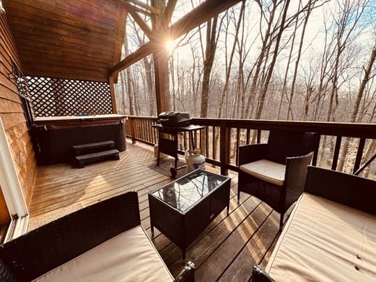A cozy deck with outdoor seating, a grill, and a hot tub surrounded by bare trees at sunset.
