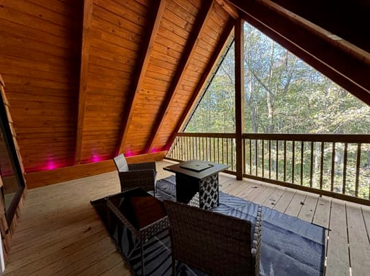 A cozy wooden deck with slanted ceilings, featuring two chairs and a small table overlooking a forested area.