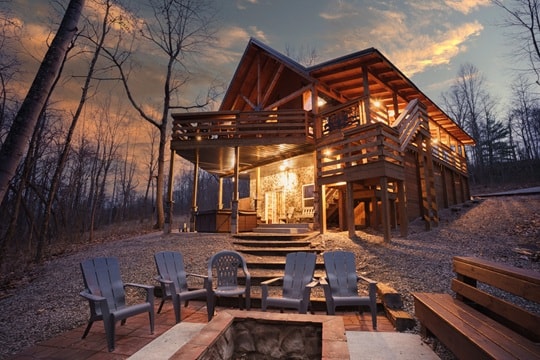 A cozy wooden cabin surrounded by trees at dusk, with a fire pit and seating area in front.