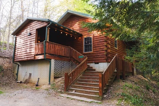 A rustic log cabin with a wooden porch and stairs, surrounded by trees.