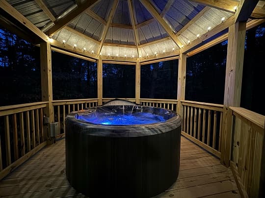 A spa tub illuminated with blue lights under a wooden gazebo at night.