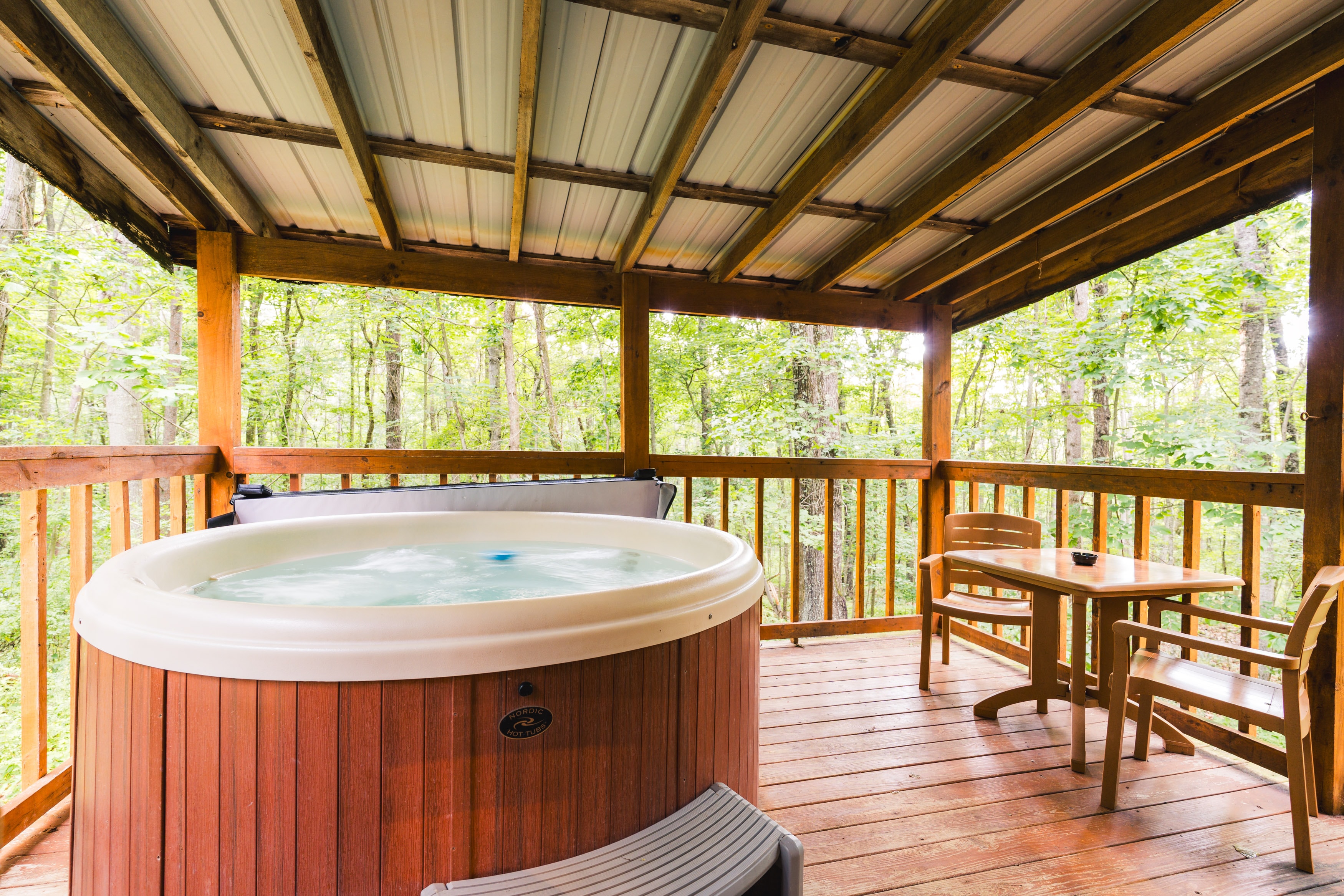 A cozy, wooden patio featuring a jacuzzi and a small table with chairs, surrounded by trees.