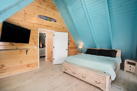 Brightly lit, modern bedroom with an A-frame ceiling, queen bed, and television against wood paneling.