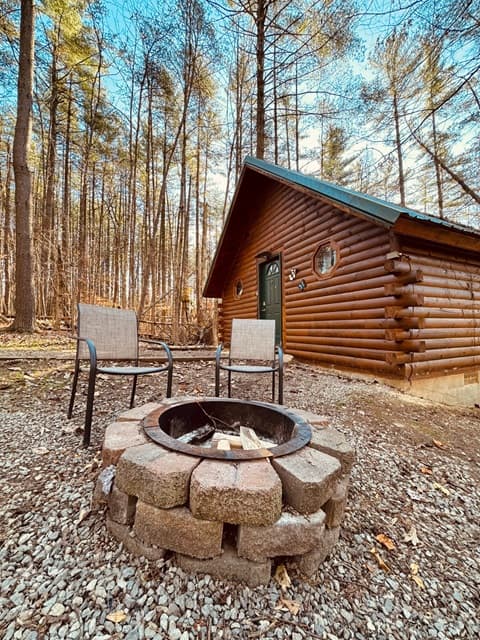 A cozy log cabin surrounded by trees, featuring a fire pit and two chairs.