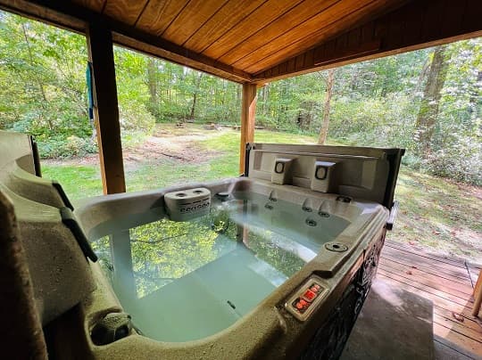 A hot tub sits under a wooden shelter, surrounded by lush greenery.