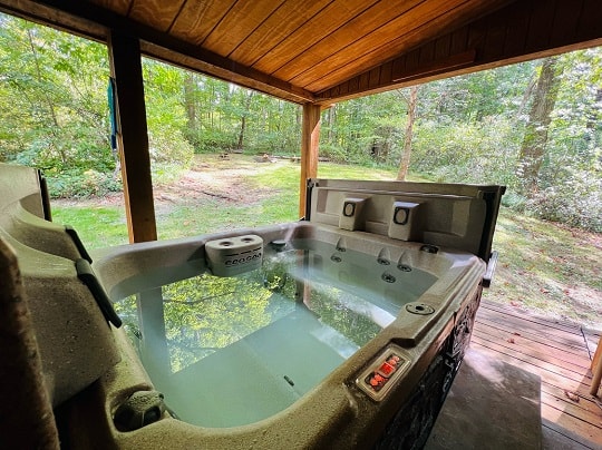 A hot tub sits under a wooden shelter, surrounded by lush greenery.