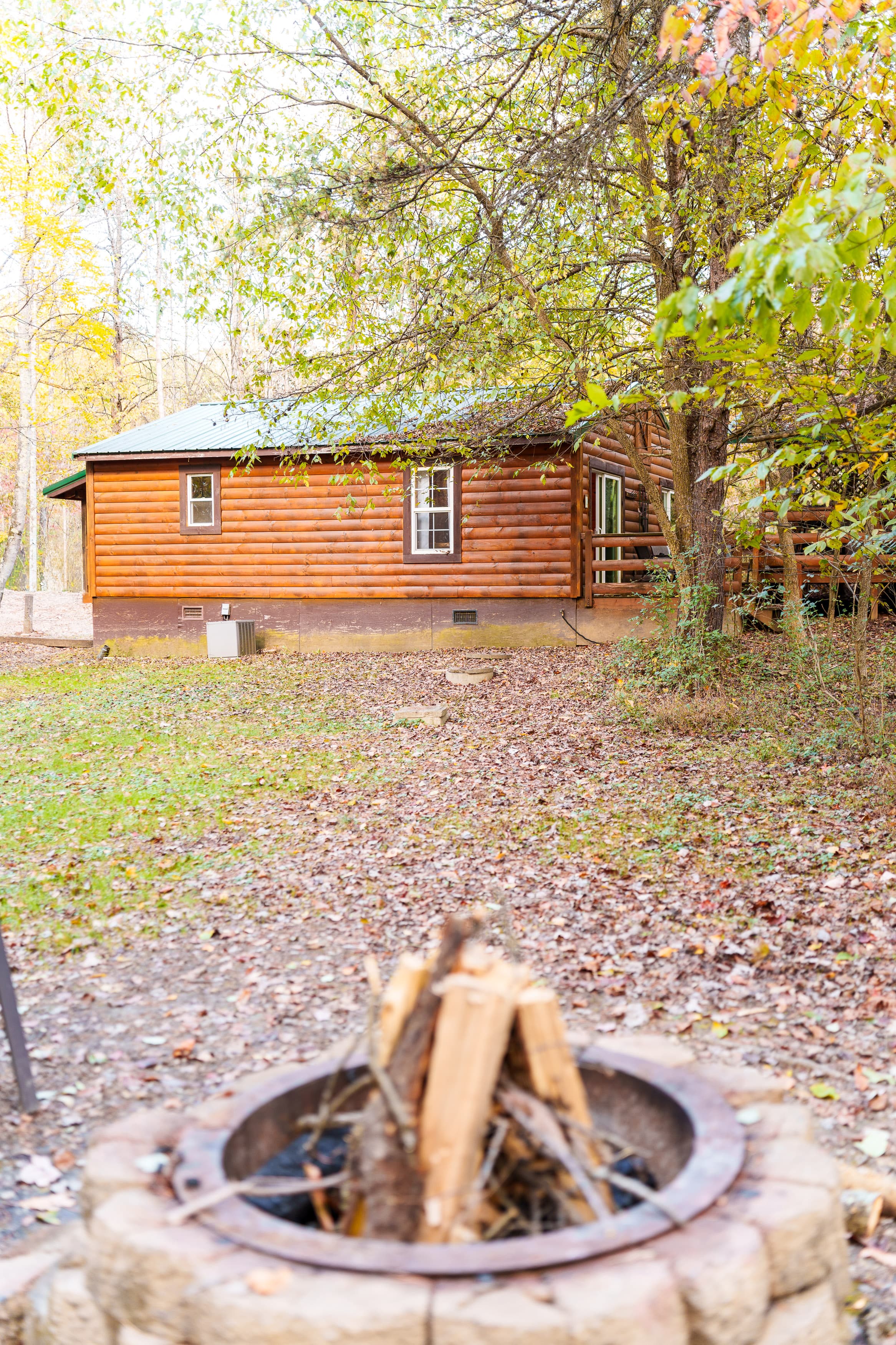A cozy log cabin surrounded by trees, with a fire pit in the foreground.