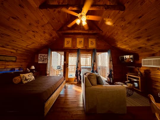 A cozy wooden cabin interior featuring a bed, sofa, and large windows leading to a porch.
