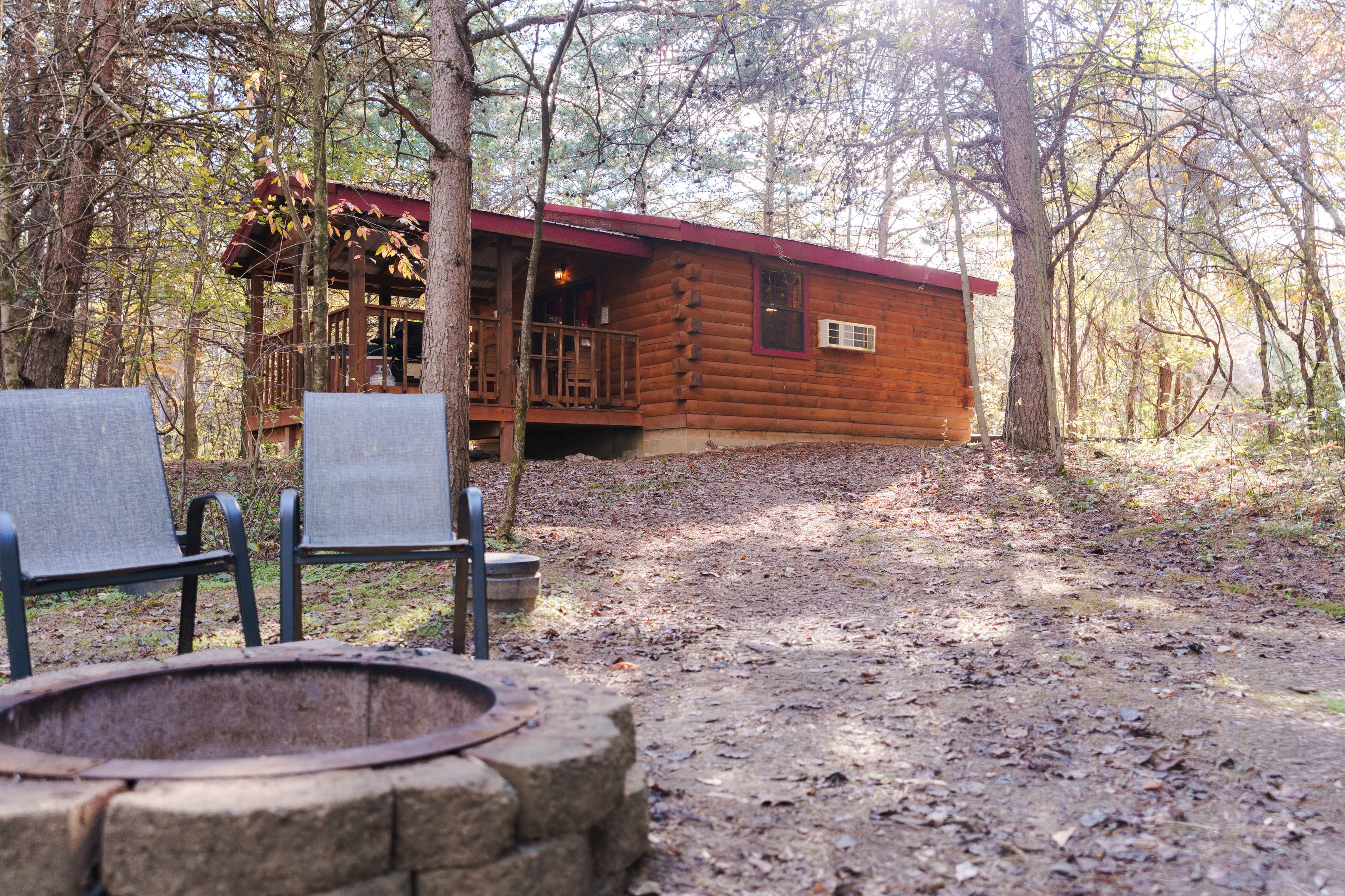 A cozy log cabin surrounded by trees, with two chairs and a fire pit in the foreground.