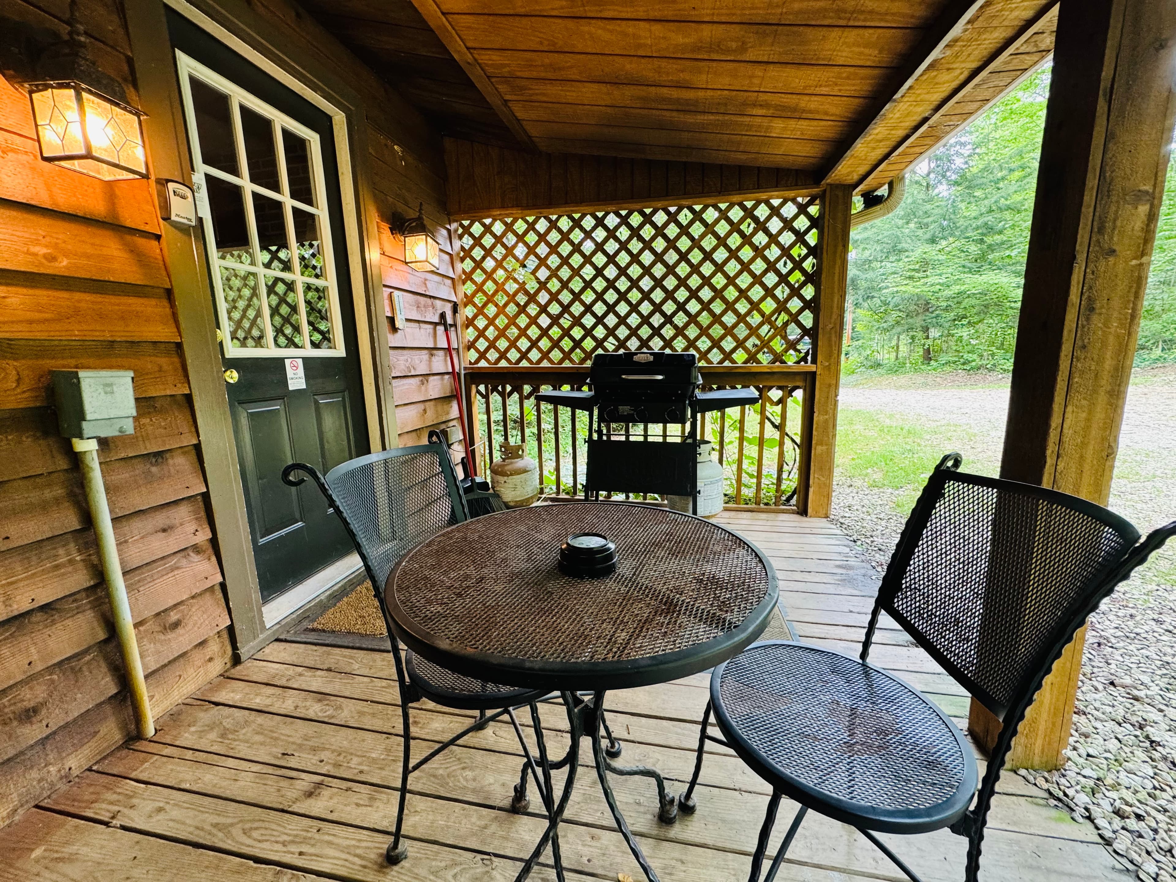 A cozy porch with a metal table and chairs, featuring a grill and surrounded by greenery.