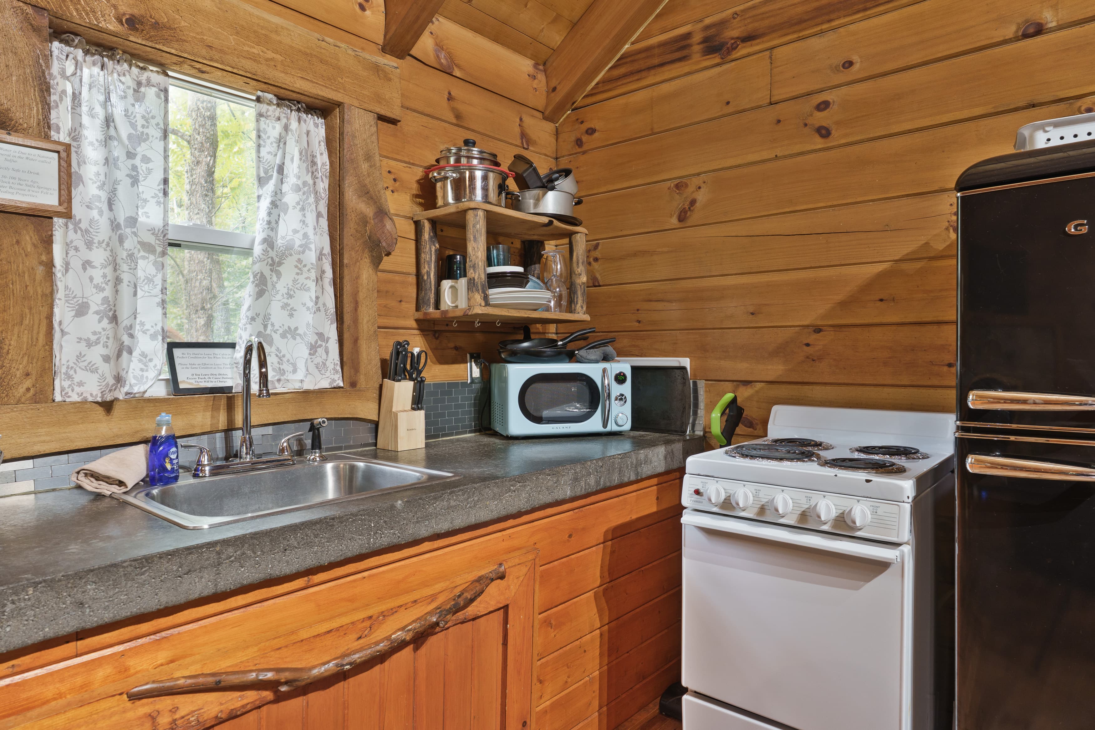 A rustic kitchen featuring wooden walls, a sink, a stovetop, a microwave, and open shelving with cooking utensils.