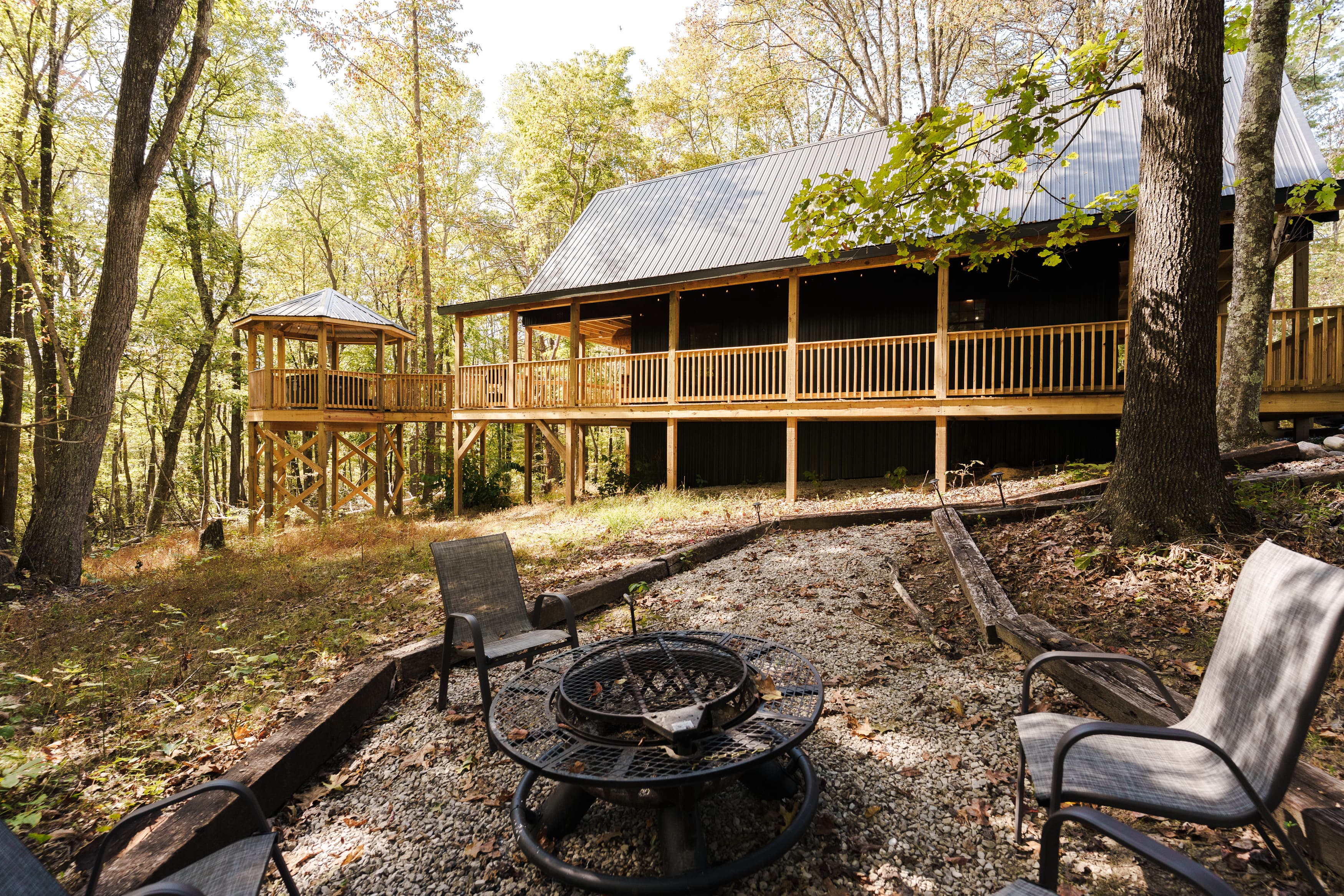 A wooden cabin with a porch overlooks a fire pit surrounded by chairs, situated in a forested area.