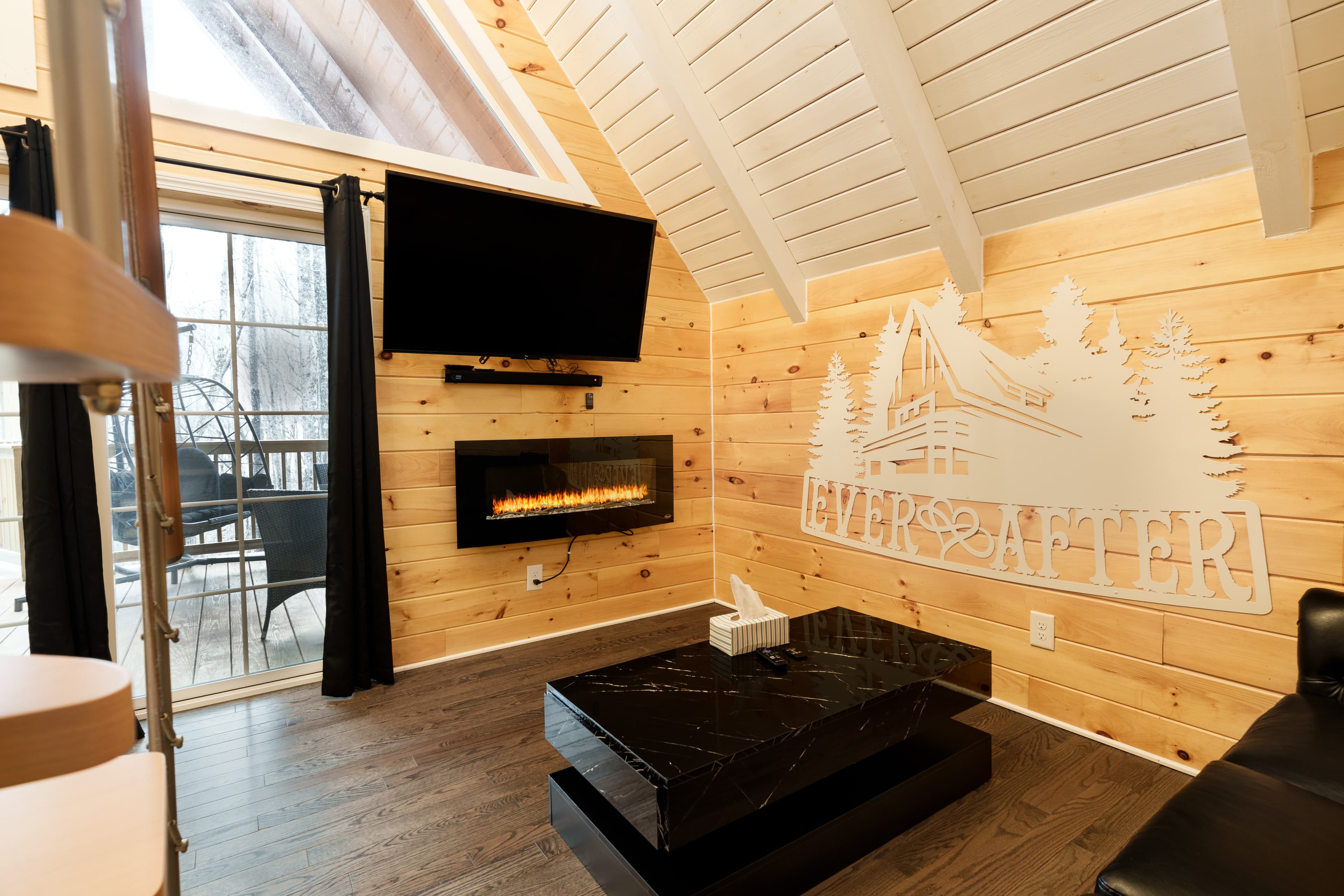 Cozy interior of a cabin featuring wooden walls, a modern fireplace, a flat-screen TV, and a sleek black coffee table.