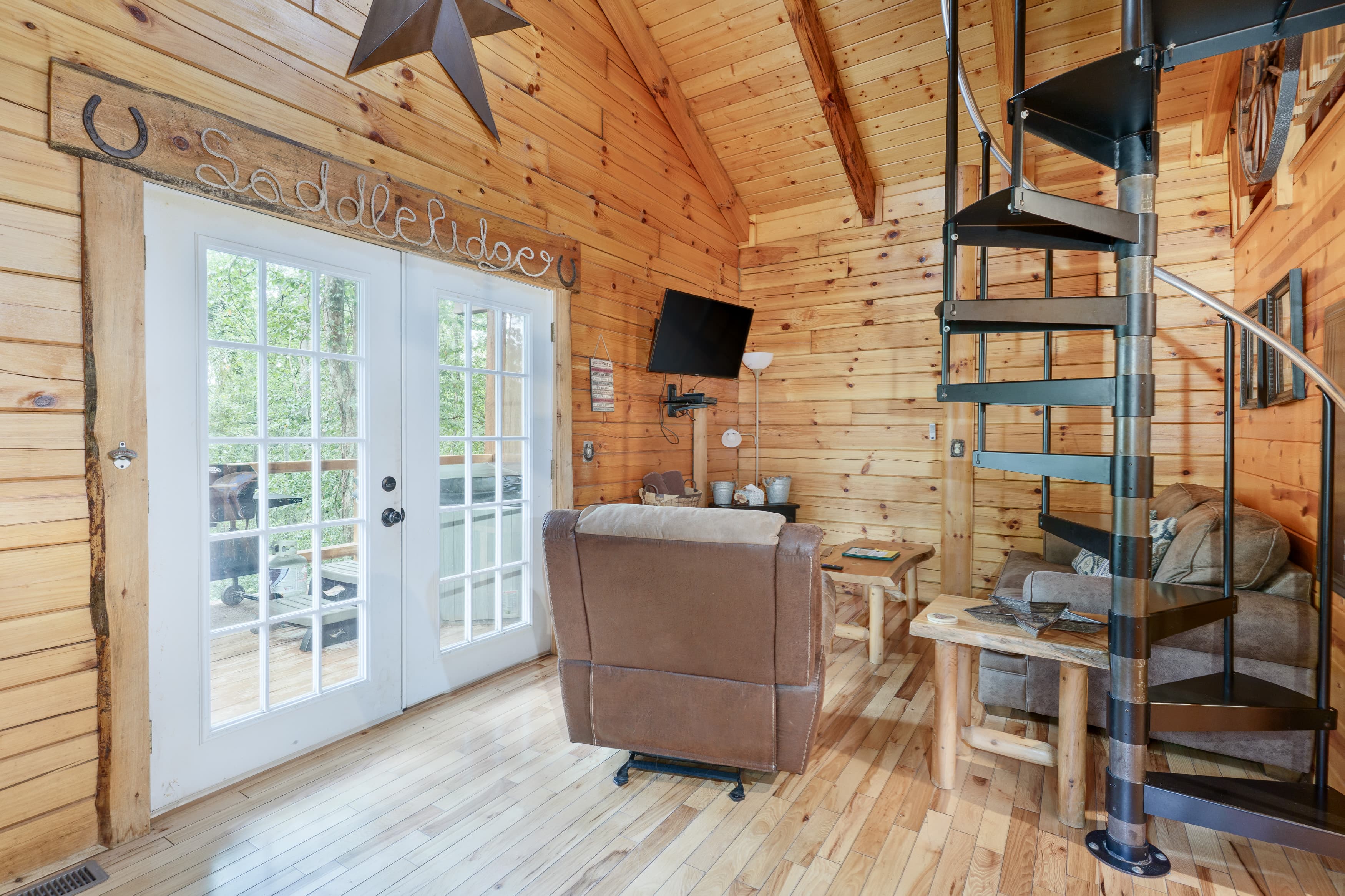 Cozy wooden interior of a cabin featuring a spiral staircase, comfortable seating, and glass doors opening to a deck.