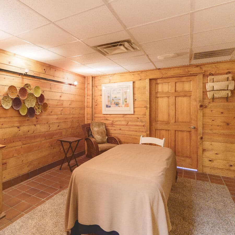 A serene, wooden-paneled room featuring a massage table, chair, and decorative wall art.