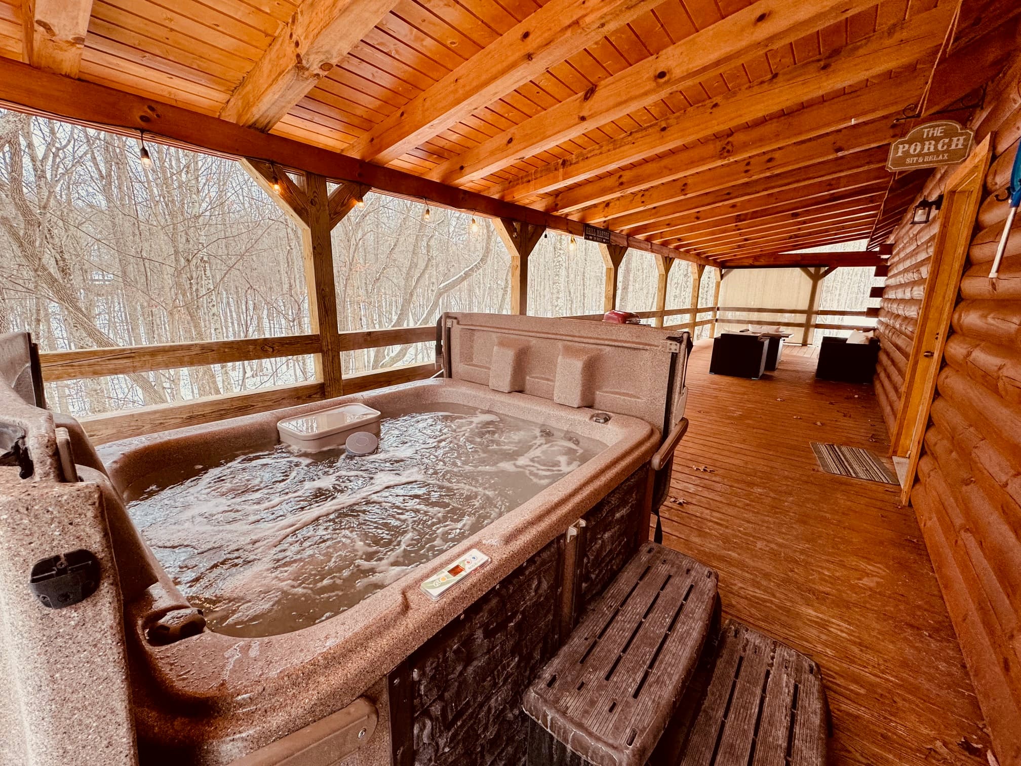 A hot tub sits on a wooden porch surrounded by trees in a cozy cabin setting.