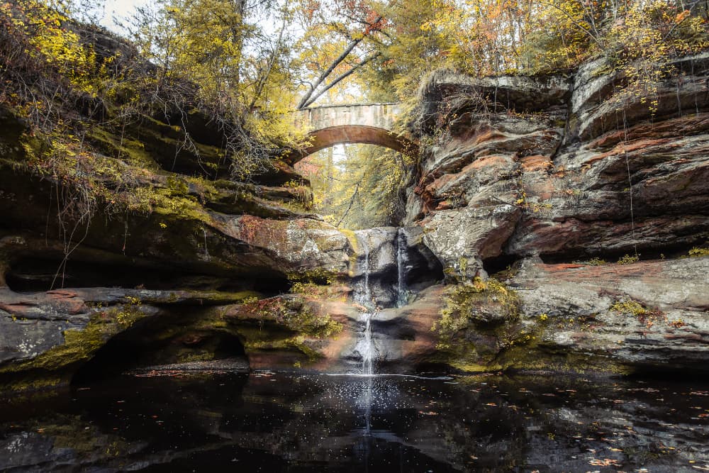 A stone bridge arches over a serene waterfall cascading into a dark pool surrounded by rocky cliffs and autumn foliage.