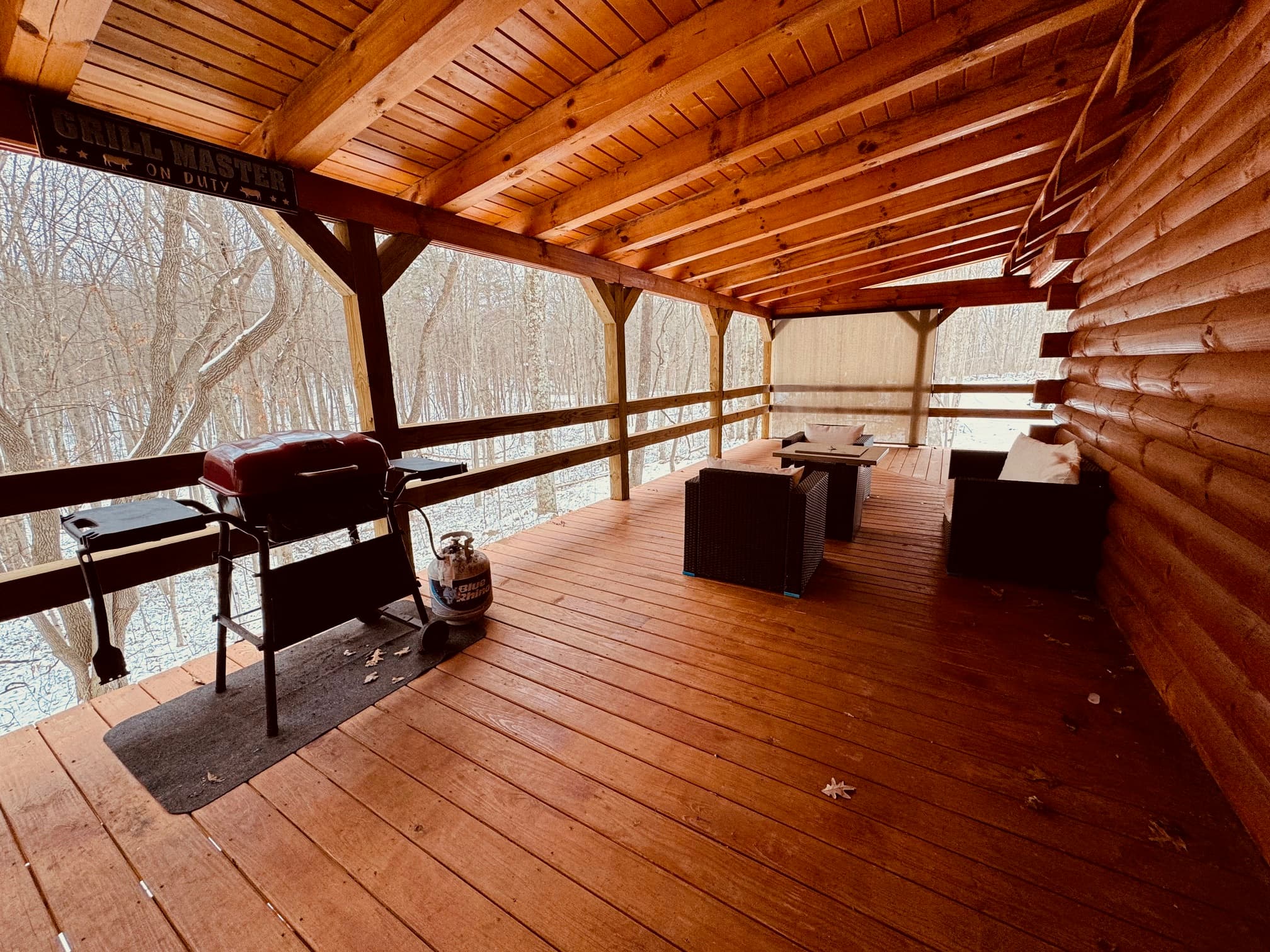 A spacious wooden porch with a grill, tables, and chairs, overlooking a snowy forest.